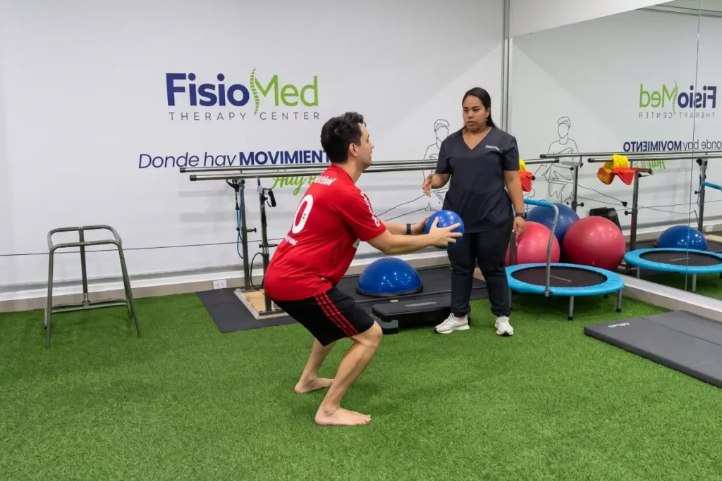 Paciente descalzo realizando una sentadilla con balón medicinal bajo supervisión de una fisioterapeuta